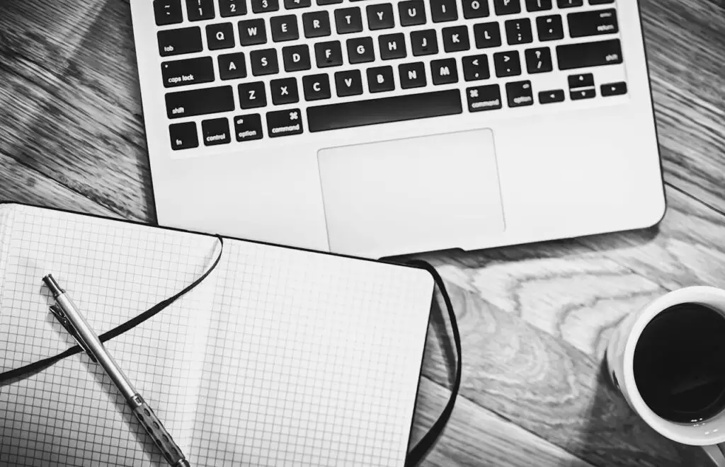 A notepad, pen, coffee, and a laptop on a wooden desk